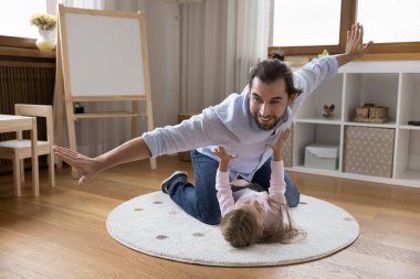 Neşeli baba, kızının dinlenen halısının üzerinde uçacak diye heyecanlı. Mutlu baba ve çocuk oyun odasında ısıtma odasında uçak oyunu oynuyorlar, gülüyorlar, eğleniyorlar, babalık, aile kavramı.