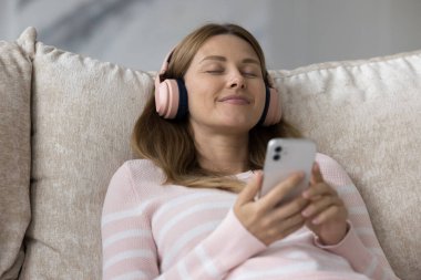 Peaceful woman in wireless headphones and cell phone in hands relaxing lying on soft sofa cushions with eyes closed listen favourite music feels carefree enjoy leisure at home using modern technology clipart