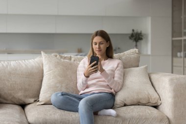 Odaklanmış akıllı telefon kullanıcısı kadın internette sohbet ediyor, cep telefonunda sanal uygulama kullanıyor, evde internet alışverişi yapıyor, dinleniyor, rahat oturuyor, keyfine bakıyor, iletişim kuruyor.