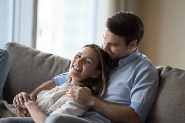 Mutlu sevgili çift rahat bir kanepede dinleniyor, şefkatle sarılıyorlar, sohbet ediyorlar, gülümsüyorlar, gülüyorlar, romantik eğleniyorlar, birlikte evde romantizmden keyif alıyorlar.,