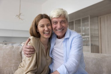 Neşeli gri saçlı yaşlı baba ve mutlu yetişkin kız evden video konferans görüşmesinde konuşuyor, dişlek gülümsemeli kameraya bakıyor, gülüyor, kanepede oturuyor. Vesikalık resmi göster