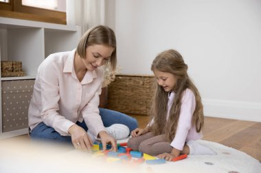 Sarışın anne ve küçük güzel kız birlikte oynuyorlar, modern çocuk odasında sıcak zeminde oturuyorlar tahta tuğlalar kullanıyorlar oyun zamanı ve iletişimin tadını çıkarıyorlar hafta sonları evde vakit geçiriyorlar.