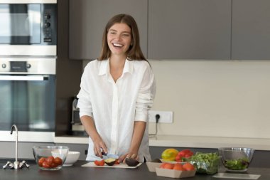 Neşeli, güzel aşçı kız akşam yemeği için sağlıklı yemekler yapıyor, mutfakta taze sebze doğruyor, salata hazırlıyor, organik gıda malzemelerini kullanıyor, uzağa bakıyor, gülümsüyor, gülüyor, konuşuyor.