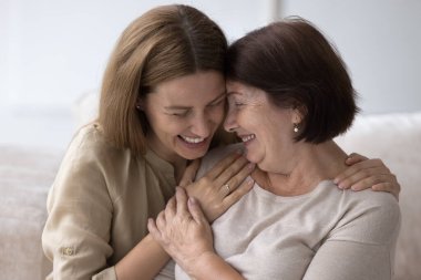 Neşeli kıdemli anne ve yetişkin kız kucaklaşıyorlar, sohbet ediyorlar, evde gülüyorlar, anneler gününü kutluyorlar. Genç bir kadın, yaşlı bir anneyi kucaklıyor gözleri kapalı, yüzüne dokunuyor.