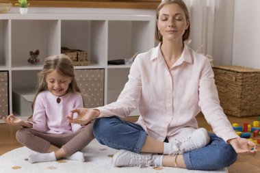 Neşeli küçük kız çocuğu ve sakin genç anne birlikte meditasyon yapıyor, ısıtma katında gözleri kapalı oturuyor, zen parmaklar yapıyor, kıkırdıyor, gülümsüyor. Anne kıza yoga yapmayı öğretiyor, eğitime odaklanıyor.