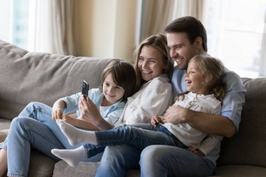 Genç anne ve baba, neşeli çocuklara kanepede sarılıyor, akıllı telefondan mutlu bir aile fotoğrafı çekiyor, cep telefonunu birlikte kullanıyor, internet iletişimi kuruyor, yerel internet teknolojisinden keyif alıyor.