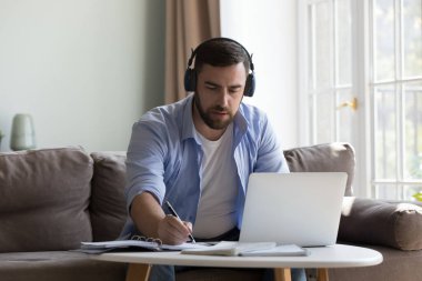 Kablosuz kulaklıklı ciddi bir milenyum adamı laptoptan video konuşması, not yazma, evdeki koltukta oturup konuşma, yetişkin bir öğrencinin online eğitim kursuna gitmesi.