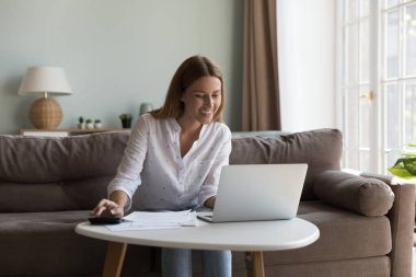Mutlu neşeli milenyum kadını kağıt dökümanlarda dizüstü bilgisayar ve hesap makinesi kullanıyor, internet üzerinden faturaları internet uygulaması ile ödüyor, monitöre bakıyor, gülümsüyor, evdeki koltukta oturuyor.