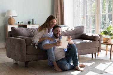 Mutlu milenyum çekici ev sahipleri evde tablet kullanıyor, kanepede dinleniyor, ısıtıyor, alışveriş yapıyor, internet uygulaması ile rezervasyon yaptırıyor, video görüşmeleri yapıyor, gülümsüyor, ekrana gülüyor.
