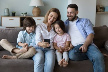 Mutlu aile kanepeye selfie çekiyor. Neşeli milenyum aileleri ve iki küçük çocuk cep telefonlarını video görüşmesi için kullanıyorlar, gülüyorlar, koltukta aletlerle rahatlıyorlar. İletişim konsepti