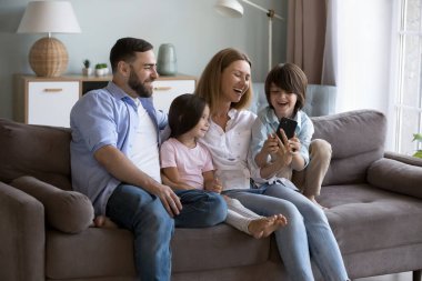 Neşeli bir çift ebeveyn ve iki çocuk birlikte yumuşak koltukta dinleniyorlar, cep telefonlarında internet servisini kullanıyorlar, internetten komik içerikler izliyorlar, video konuşmaları yapıyorlar, selfie çekiyorlar, gülüyorlar, gülüyorlar.