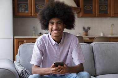 Uzun kıvırcık saçlı mutlu Afrikalı çocuk akıllı telefonu evde kullanıyor, kameraya bakıyor, gülümsüyor, gülüyor, kanepede oturuyor. Siyah, genç gezici hpne kullanıcı portresi