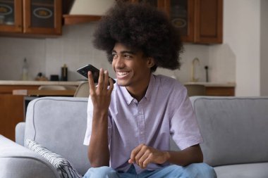 Afrika saçı olan neşeli genç adam akıllı telefondan hoparlörde konuşuyor, hayalperest, düşünceli bir gülümsemeyle, gülüyor, sesli mesaj kaydediyor, sanal asistanı kullanıyor.