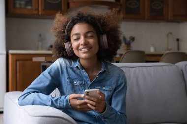 Büyük kablosuz kulaklıklı mutlu genç Afrikalı kız. Ortam müziği dinliyor, müzik listesini gevşetiyor, evdeki kanepede oturuyor, gözleri kapalı ve dişlek gülümsüyor.