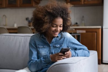 Neşeli, güzel, genç Afrikalı kız sanal uygulama kullanıyor, cep telefonunda internet servisi var, sohbet ediyor, video görüşmesi yapıyor, kanepede oturuyor, ekranda gülümsüyor, gülüyor.