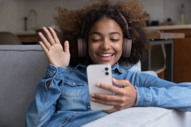 Neşeli gen Z akıllı telefon kullanıcısı. Kablosuz kulaklıklı kız. Video görüşmesiyle konuşuyor, merhaba diyor, gülümsüyor, gülüyor. Afrikalı genç cep telefonu kullanıyor, kanepede dinleniyor.