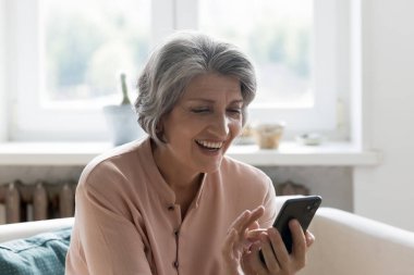 Heyecanlı mutlu cep telefonu kullanıcısı kadın görüntülü konuşma yapıyor, iyi haberlerle mesaj okuyor, gülüyor. Son sınıf gri saçlı bayan internet uygulamasını cep telefonunda kullanıyor, alışveriş yapıyor, indirim yapıyor, kar elde ediyor.