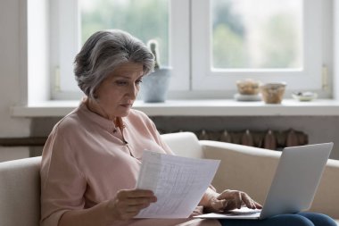 Ciddi, olgun muhasebeci kadın laptopta ödeme kağıtlarını kontrol ediyor, faturaları, faturaları ödüyor, sigorta ücretlerini ödüyor, bütçe sayıyor, giderleri buluyor, zarar ediyor, maddi sorunlar yaşıyor.