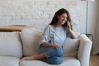 Neşeli, mutlu, genç, İspanyol bir kadın cep telefonuyla konuşuyor, telefonla konuşuyor, telefon ediyor, komik haberleri paylaşıyor, evdeki kanepede dinleniyor, gülüyor, gülümsüyor. İletişim konsepti