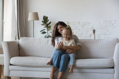 Neşeli anne ve küçük çocuk görüntülü konuşuyorlar. Mutlu blogcu anne kucağında güzel bir kız çocuğu, evinde kanepede oturup selfie çekiyor. Annelik, iletişim konsepti