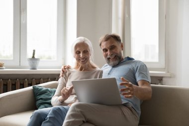 Mutlu, heyecanlı, olgun bir çift evde dizüstü bilgisayar kullanıyor, kanepeden video görüşmesi yapıyor, konuşuyor, gülüyor, ekranda gülümsüyor. Mutlu kıdemli karı koca online uygulama kullanarak iyi haberler alıyorlar.