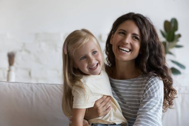 Mutlu, genç bir anne, güzel küçük kızını kucaklıyor, kucağında çocuğu sevgiyle kucaklıyor, şefkatle kucaklıyor, dişlek tebessümle başka tarafa bakıyor, gülüyor, poz veriyor, anneliğin tadını çıkarıyor.