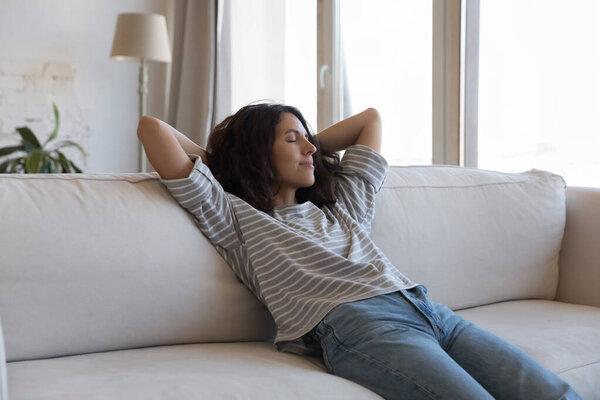 Calm sleepy pretty young Hispanic woman resting on soft couch, leaning on back, breathing fresh pure air, relaxing, enjoying leisure, practicing meditation, stress relief with closed eyes at home