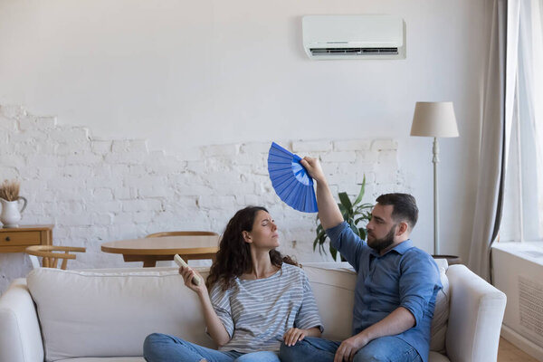 Young couple frustrated with heat, waving handheld fan, using remote control, turning air conditioner on for cooling air, sitting on couch at home. Domestic equipment failure concept