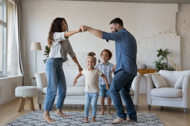 Mutlu genç ebeveynler ve küçük çocuklar birlikte aktif oyunlar oynuyorlar, oturma odasında müzik eşliğinde dans öğreniyorlar, el ele tutuşuyorlar, eğleniyorlar, taşınmayı kutluyorlar, ev satın alıyorlar, eğleniyorlar.