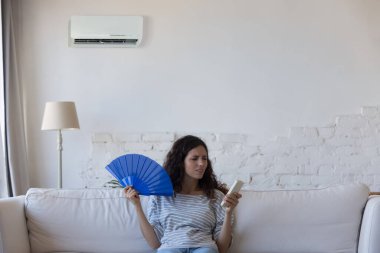 Kızgın ev sahibi genç kadın sıcaktan usanmış, kendini rahatsız hissediyor, soğutma vantilatörü sallıyor, bozuk klima için uzaktan kumanda tutuyor, evdeki kanepede oturuyor.