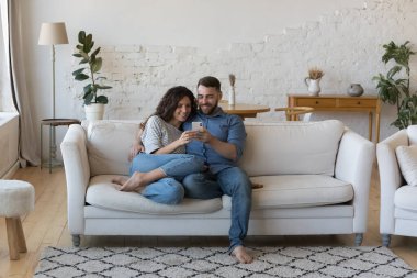 Mutlu milenyum karı ve koca akıllı telefonu evde birlikte kullanıyor, cep telefonunu paylaşıyor, internetten online ekommerce uygulaması ile alışveriş yapıyor, video görüşmeleri yapıyor, kanepede dinleniyor, sarılıyor, gülümsüyor.