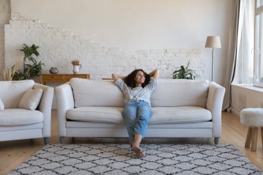 Mutlu düşünceli genç Latin kadın evdeki rahat kanepede dinleniyor, başka tarafa bakıyor, gülümsüyor, hayal kuruyor, düşünüyor, dinleniyor, eğleniyor, temiz hava alıyor.