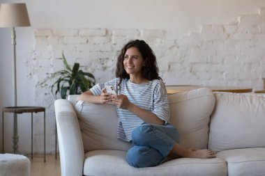 Neşeli, hayalperest, akıllı telefon kullanıcısı kadın, evde oturuyor, başka tarafa bakıyor, mutlu düşüncelere, fikirlere, planlara, daktilo yazma mesajlarına gülüyor, internetten sohbet ediyor, düşünüyor, akıllı telefon tutuyor.