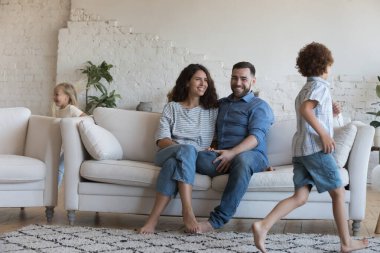 Mutlu ebeveynler, kanepede koşuşturan, kovalayan, oturma odasında aktif oyunlar oynayan iki hiperaktif küçük kardeş çocuğu izliyor. Çift ve çocuklar, aile eğlencesi, oyun zamanı.