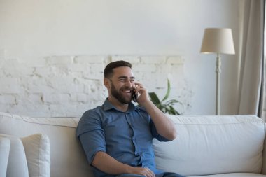 Neşeli milenyum adamı evde cep telefonuyla konuşuyor, telefonla konuşuyor, konuşuyor, iyi haberleri dinliyor, eğleniyor, iletişim kuruyor, gülüyor, kanepede dinleniyor.