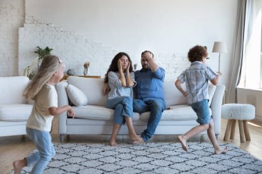 Yorgun ebeveynlerin etrafında koşuşturan iki aktif kardeş çocuk, aktif oyunlar oynuyorlar. Annem ve babam çocuklara bakmaktan yoruldular, baş ağrısı, stres hissettiler. Yetiştirme sorunları, ebeveynlik, aile kavramı