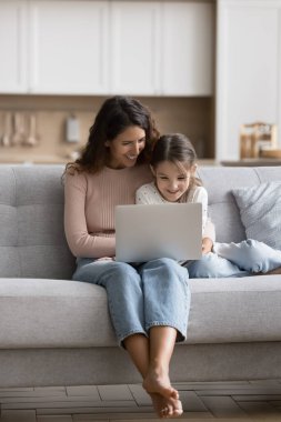 Dikey çekim genç anne ve küçük kızı online filmler izlerken modern dizüstü bilgisayarla koltukta oturuyor, ürünleri ve e-hizmetleri seçiyor, ilgileniyor gibi görünüyor, evde internette boş zaman geçiriyor. Teknoloji