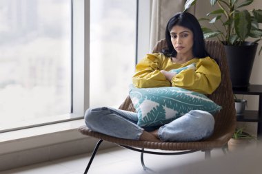 Bunalımlı genç Hintli kadın evinde koltukta oturuyor, yastığı sıkıyor, boşluğa bakıyor, başka tarafa bakıyor, sorunları düşünüyor, başarısızlık, kötü haber, kayıp, depresyondan muzdarip