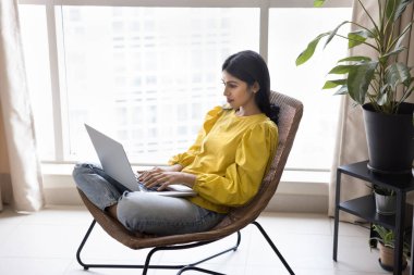 Ciddi Hintli serbest çalışan kız rahat bir şezlongda çalışıyor, dizüstü bilgisayarla yazı yazıyor, penceredeki koltukta oturuyor, bilgisayarla uzaktan kumandayla iletişim kuruyor.