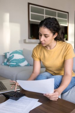 Ciddi bir Latin serbest muhasebeci evde mali muhasebe evrakları için hesap makinesi kullanıyor, kanepede oturuyor, hesap yapıyor, ipotek taksitlerini hesaplıyor, fıçı, vergiler, kağıt döküman tutuyor. Dikey çekim