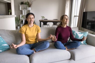 Pozitif kıdemli İspanyol anne ve huzurlu genç kız evde gözleri kapalı meditasyon yapıyor, gözleri kapalı gülümsüyor, kanepeye oturmuş meditasyon yapıyor, zen yapıyor, el hareketlerini sakince yapıyor.
