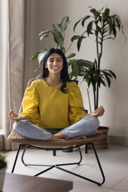 Neşeli genç Hintli kadın evdeki şezlongda meditasyon yapıyor, zen parmaklarını kapalı tutuyor, gülümsüyor, gülüyor, temiz hava soluyor, stres atağı yapıyor, konsantre oluyor.
