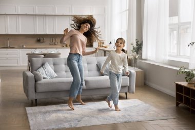 Rahat giyinen, eğlenen, eğlenen, 6 'lı yaşlardaki kızıyla dans eden, hafta sonu boş vakitlerinde sıcak oturma odasında çıplak ayakla halının üzerinde zıplayan kaygısız bir kadın. Etkin eğlence