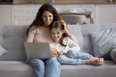 Genç anne ve küçük kızı birlikte kanepede oturup dizüstü bilgisayara bakıyorlar, aile filmi izlemekten, fotoğraflara bakmaktan, çevrimiçi oyun oynamaktan, online eğitim aktivitelerine katılmaktan zevk alıyorlar.