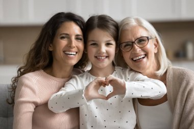 Sevimli 6 'lı yaşlardaki küçük bir kızın portresi birleşik parmaklarla aşk sembolü gösteriyor, yaşlı büyükanne ve genç anneyle kameraya poz veriyor. Güçlü aile bağları, nesiller boyu sevgi ve duygusal bağ, özen.