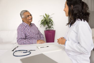 Gülümseyen Hintli kıdemli bir adam klinikte genç Brezilyalı kadın kardiyologla konuşuyor, hastalığın etkili tedavisi hakkında iyi haberler alıyor, profesyonel sağlık hizmetlerini tatmin ediyor.