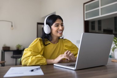 Kablosuz kulaklıklı Hintli pozitif öğrenci dizüstü bilgisayarı izleyip evde internetten ders çalışıyor. Serbest çalışan iş kadını video konuşmasında konuşuyor, iş için müzik dinliyor.