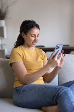 Evde aletlerle eğlenen mutlu, genç İspanyol kız cep telefonuyla akıllı ev uygulaması kullanıyor, evdeki kanepede dinleniyor, internette geziniyor, sosyal medyada gülümsüyor. Dikey çekim