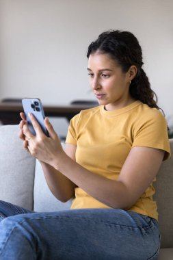 Güzel genç 20 'li yaşlarda Latin bir kadın evde akıllı telefonuyla ev uygulaması kullanıyor. Dijital aletlerle koltukta dinleniyor, internet video içeriğini izliyor, internet iletişiminden keyif alıyor.