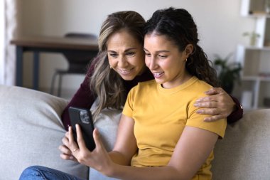 Mutlu kıdemli İspanyol anne ve yetişkin kız sosyal medyada birlikte geziniyor, online teknoloji kullanıyor, cep telefonunda uygulama yapıyor, internet içeriğini izliyor.